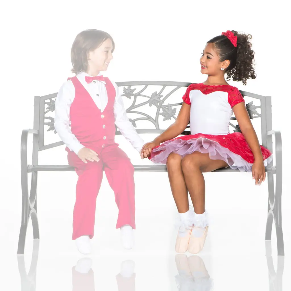 Two children in red formal outfits sit holding hands on bench.