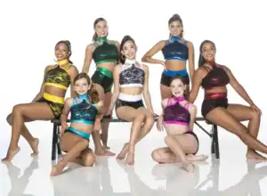 Seven dancers pose in colorful costumes on a bench.