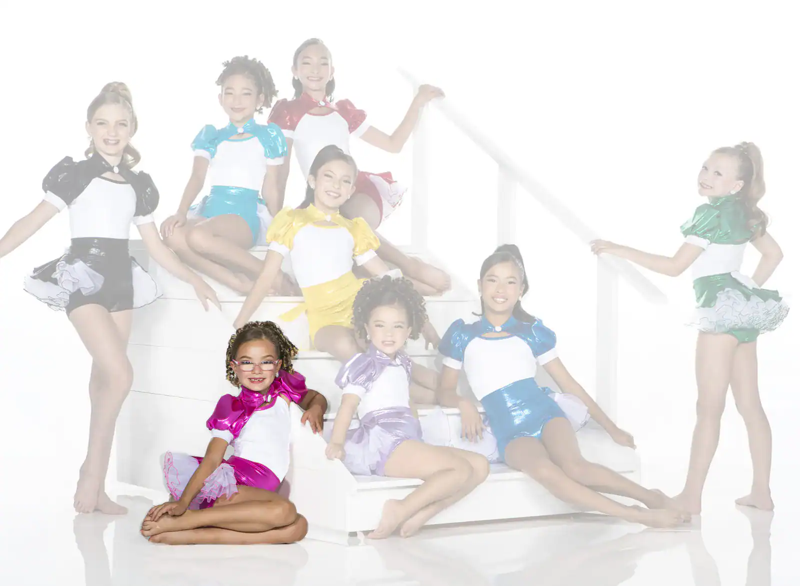 Seven young dancers in colorful costumes pose on white stairs.