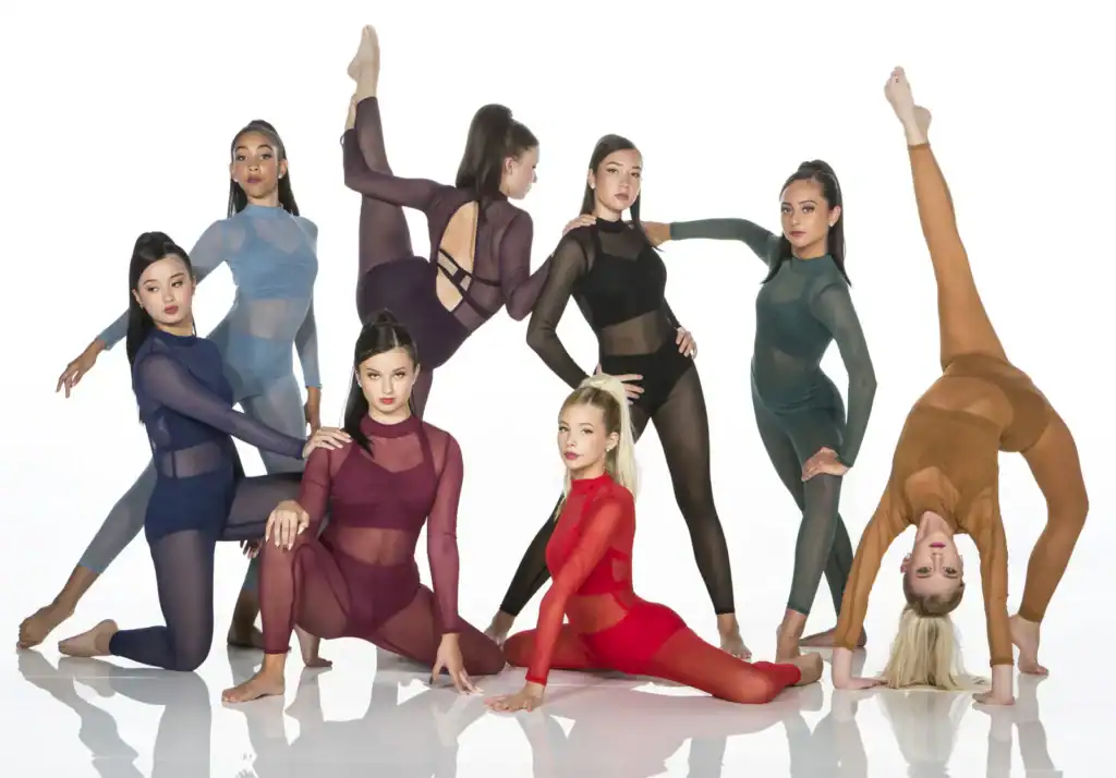 Seven dancers pose in colorful sheer leotards on white background.