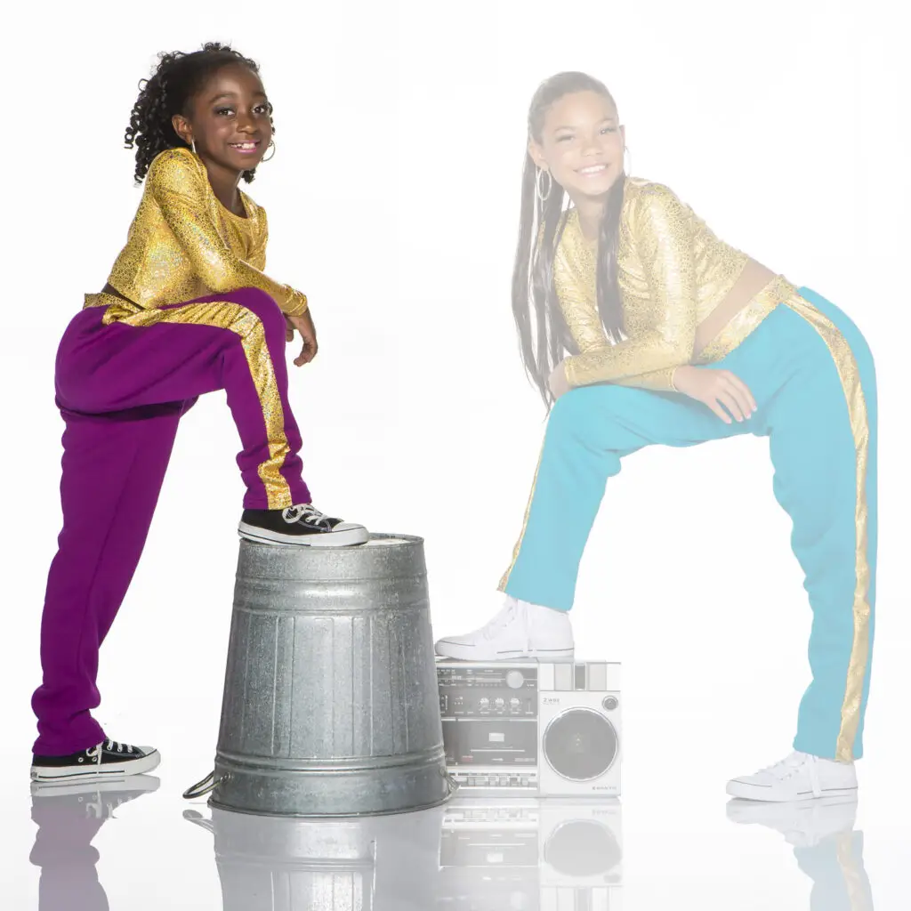 Two girls in glitter outfits pose with boombox and trash can.