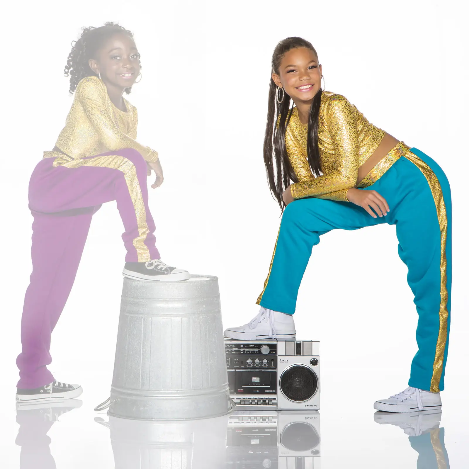 Two girls in gold tops pose beside boombox and trash can.